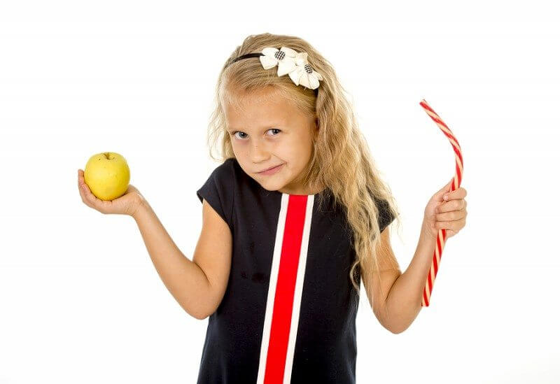 Motivation and autism: little beautiful female child with blond hair choosing dessert holding unhealthy but tasty red candy licorice and apple fruit in healthy versus unhealthy die nutrition isolated on white background